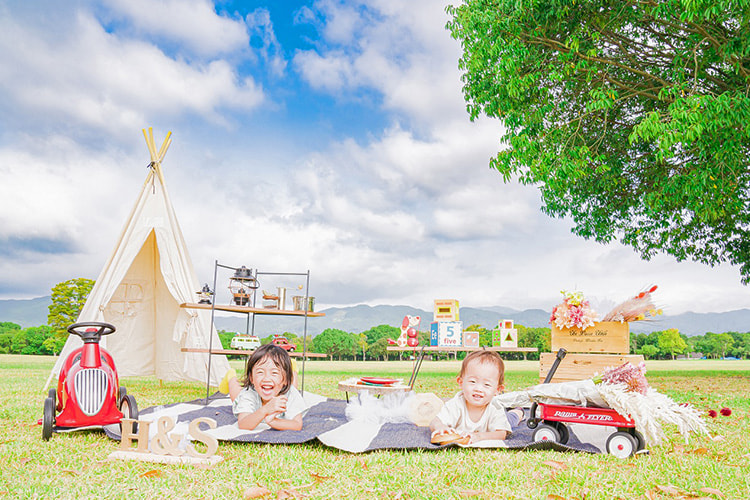 芝生広場に置かれたテントやおもちゃを背景に、芝生に寝転がる2人の子どもの写真