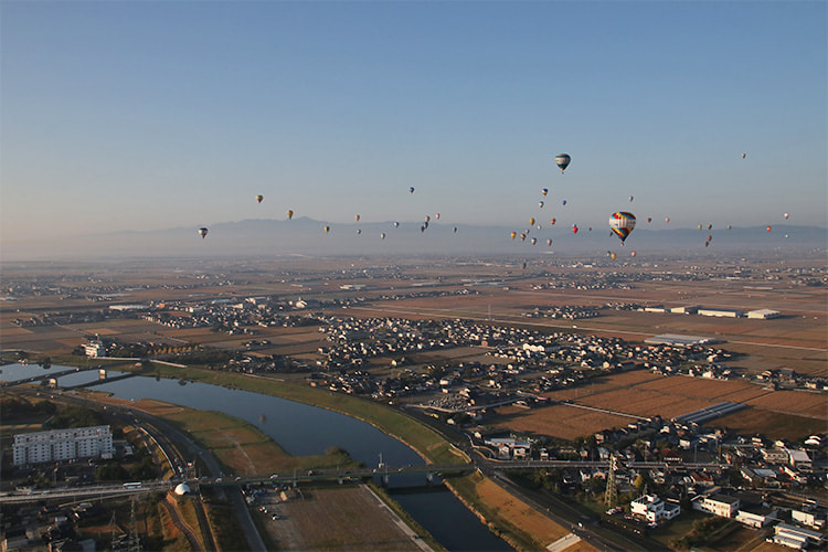 離陸した河川敷から西へと向かう熱気球群の写真