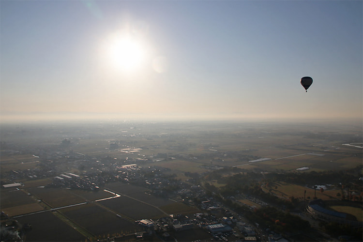 遠くまで続く朝もやの地上を照らす太陽とシルエットの熱気球の写真